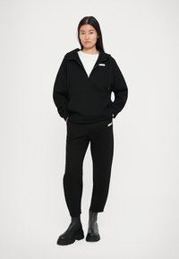 Črna zip-up trenerka s sprednjim žepom in kapuco z vrvico, v kombinaciji s črnimi ozkimi hlačami in robustnimi črnimi škornji. Gladka tekstura.