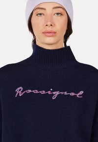 Navy knitted sweater with high collar, featuring multicolored embroidered "Rossignol" logo across the chest. Soft texture, ribbed detailing.
