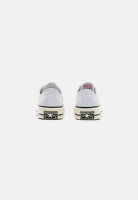 Valged kangast tenniset kurviga pinnaga, madala profiiliga disain, tume talla ja ikoonilise musta ja valge Converse All Star brändinguga kontsaplaanil.