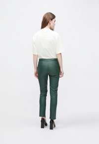 Groene leren broek met een aansluitend ontwerp, gecombineerd met een lichtgekleurde t-shirt. Model gezien van achteren, met zwarte blokhakken.