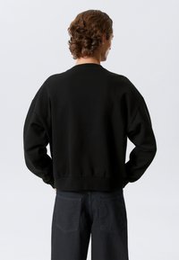 Juodas džemperis laisvo silueto, su ribotais rankogaliais ir apačia. Minkšta medžiaga su vienspalve tekstūra. Dėvimas su tamsiais džinso kelnėmis.