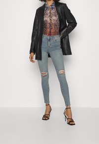 Zwart leren blazer, multicolore slangenprint shirt, lichtblauwe skinny jeans met scheuren, en zwarte gekruiste sandalen met hoge hakken.