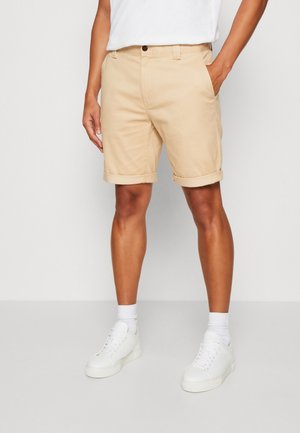 Homme portant un short beige retroussé, des chaussettes blanches, des baskets blanches et une chemise blanche, une main dans la poche, debout sur un sol gris clair.