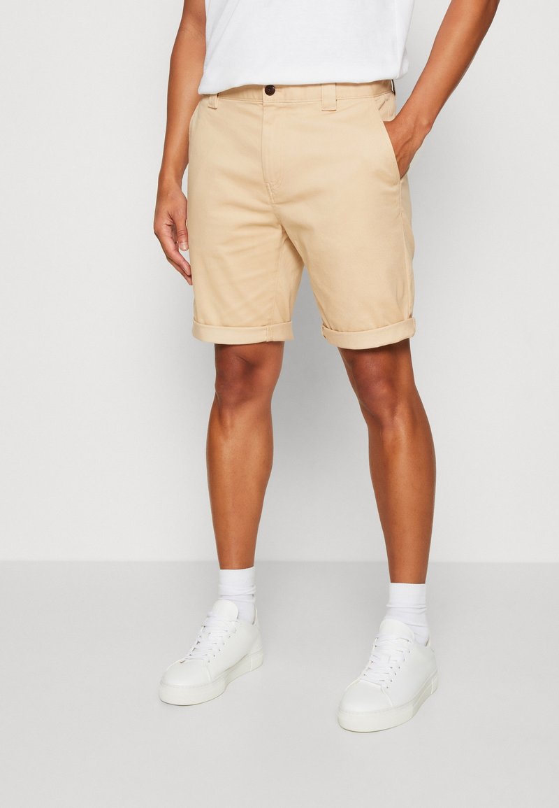 Homme portant un short beige retroussé, des chaussettes blanches, des baskets blanches et une chemise blanche, une main dans la poche, debout sur un sol gris clair.