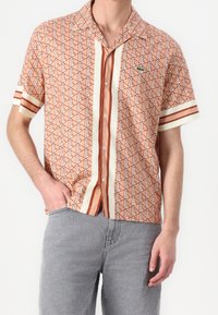 Chemise à manches courtes de couleur pêche et crème avec des motifs géométriques, dotée d'un col et d'un devant à boutons. Comprend des accents rayés sur les manches.