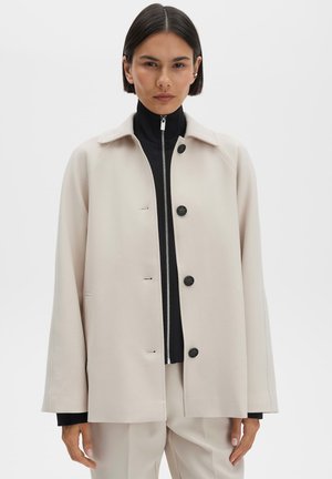 Beiger Mantel aus Wollmischung mit lässiger Passform, schwarzen Knöpfen und aufgesetzten Taschen. Getragen über einem schwarzen Reißverschlussoberteil mit langen Ärmeln.