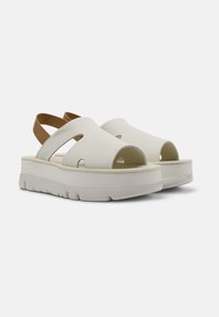 White platform sandals with open toe and heel, featuring cut-out designs and a contrasting brown ankle strap. Soft material and textured sole.