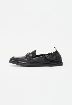 Mocasín de cuero negro con parte trasera elástica, suela plana baja, hebilla redonda de metal en la parte superior y tira para tirar en el talón.