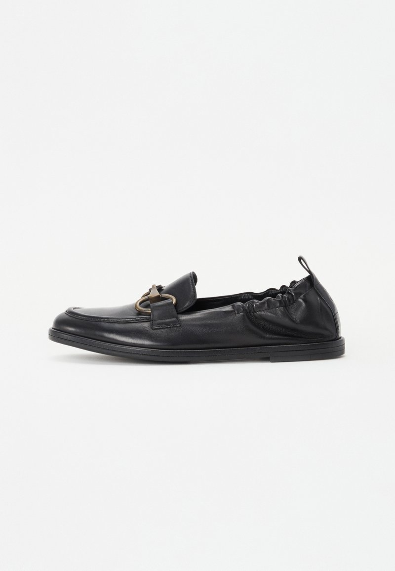 Mocassin en cuir noir avec arrière élastiqué, semelle plate basse, boucle ronde en métal sur le dessus et languette à l'arrière.