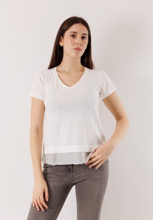 Giovane donna con lunghi capelli scuri indossa una maglietta bianca a maniche corte a strati e jeans grigi, in piedi davanti a uno sfondo chiaro e semplice.