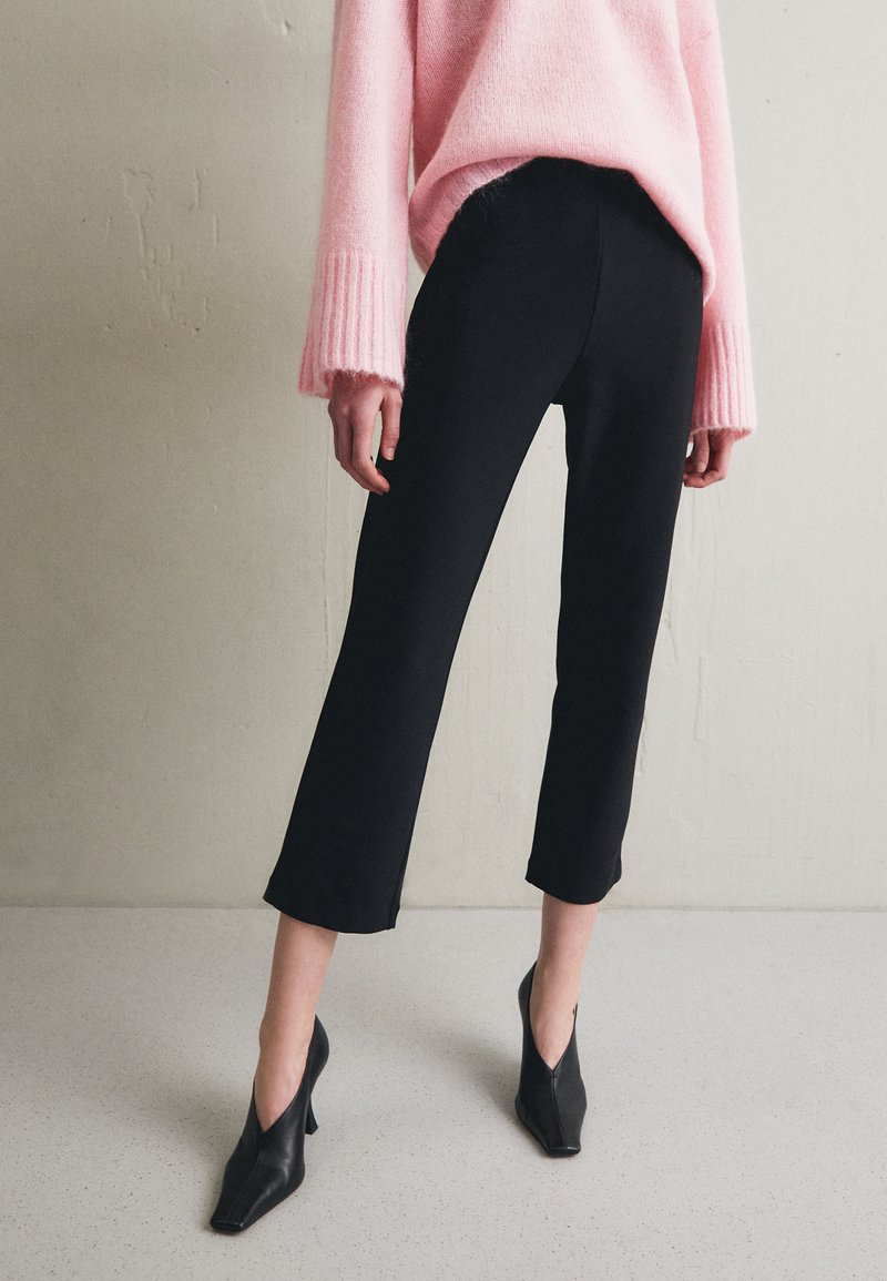 Black tailored pants with a cropped, straight-leg cut, paired with a soft, light pink sweater. The shoes are black, open-toe heels.