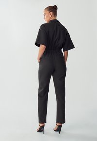 Zwarte jumpsuit met korte mouwen, elastische tailleband en taps toelopende benen. Glad textuur met een minimalistisch ontwerp. Draagt met hoge hakken sandalen.