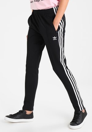 Osoba nosząca czarne spodnie dresowe Adidas z białymi paskami i logo, w połączeniu z czarnymi trampkami i różową koszulką.