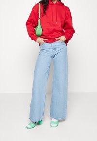 Czerwona bluzka z kapturem, jasnoniebieskie szerokie dżinsy, zielona torebka i zielone sandały na obcasie z białymi akcentami. Gładkie białe tło.