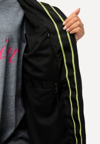 Svart jacka med nättextur på insidan, som har två vertikala gröna ränder och en dragsko-ficka. Grå skjorta är delvis synlig.