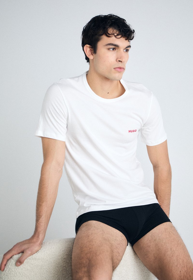 T-shirt en coton blanc à manches courtes avec un logo rouge "HUGO", associé à un caleçon noir. Le modèle pose assis sur une surface texturée.