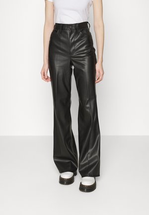 Osoba ubrana w czarne skórzane spodnie o szerokich nogawkach z wysokim stanem, białą koszulkę i białe buty na platformie, stojąca na tle jednolitego tła.