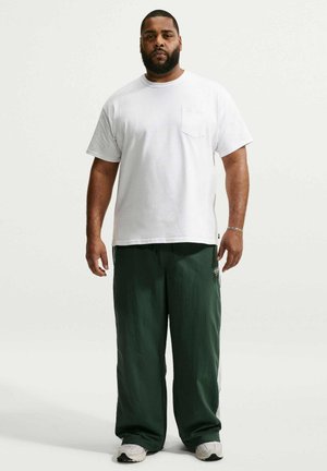 Mand med skæg står vendt fremad iført en hvid T-shirt med korte ærmer, mørkegrønne løstsiddende bukser, hvide sneakers og et sølvarmbånd.