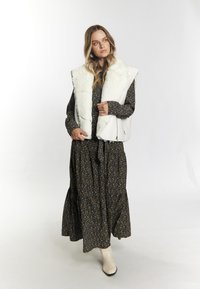 Witte faux fur vest over een donker bloemenlang jurkje. Het jurkje heeft een gelaagd ontwerp en het model draagt lichtgekleurde enkellaarsjes.