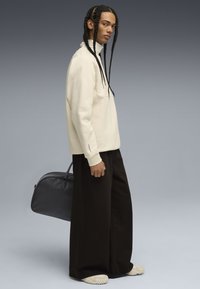 Felpa beige, taglio rilassato, colletto alto, abbinata a pantaloni larghi neri e scarpe crema. Una borsa nera elegante è tenuta in una mano.