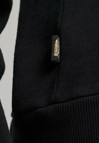Tejido de sudadera negro con una cintura acanalada. Presenta una pequeña etiqueta en el lateral con un diseño de acento dorado. Textura suave.