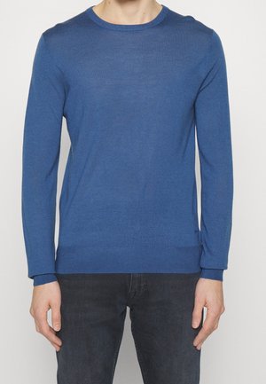 Homme portant un pull bleu uni à manches longues et un jean foncé, debout sur un fond blanc.