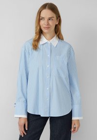 Lichtblauw gestreept shirt met een witte kraag en manchetten. Heeft een knoopsluiting aan de voorkant en een borstzak aan de linkerkant. Losse pasvorm met lange mouwen.