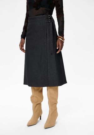 Vrouw in een zwarte midi wikkelrok met dubbele gespstraps, een doorzichtige zwarte bloeiende blouse met lange mouwen, tan gekleurde suède hoge hakken en armbanden.
