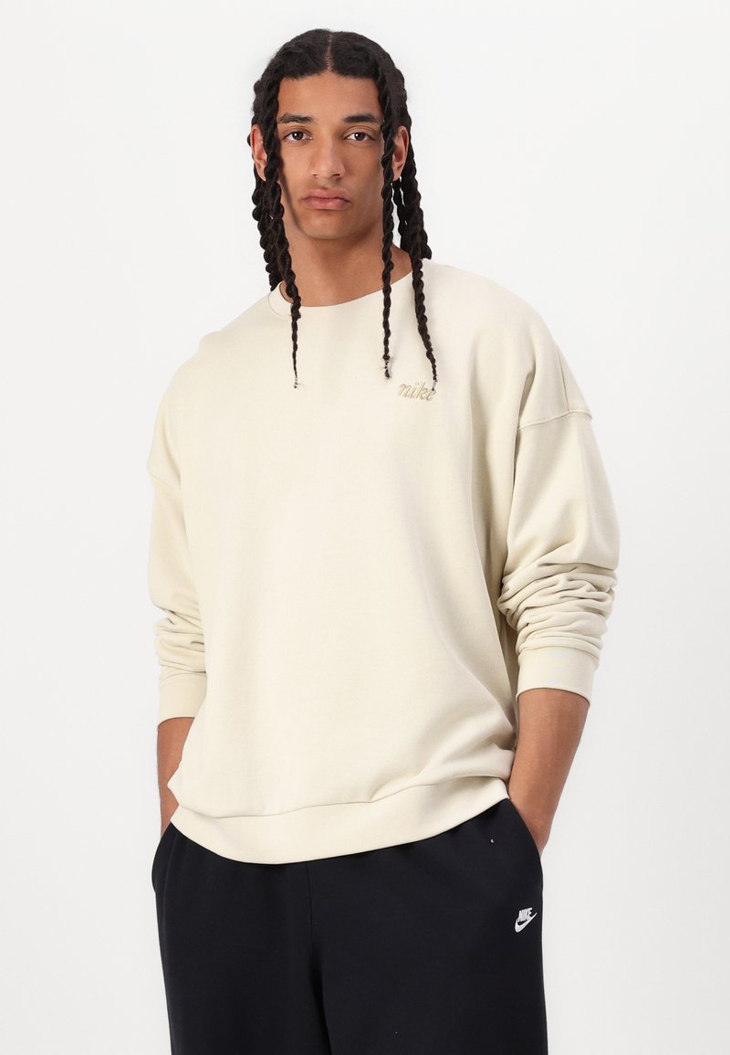 Sweat-shirt Nike couleur crème avec une coupe décontractée, des poignets côtelés et un petit logo brodé sur la poitrine. Porté avec un pantalon de survêtement noir.