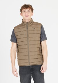 Gevoerde vest in lichtbruin met een hoge kraag, voorzien van horizontale quiltlijnen, een ritssluiting aan de voorkant en zijzakken. Grijs T-shirt eronder.