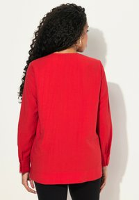 Maglietta rossa a maniche lunghe realizzata in tessuto texturizzato con una linea dritta, caratterizzata da un collo rotondo e polsini con bottoni, indossata con pantaloni neri.