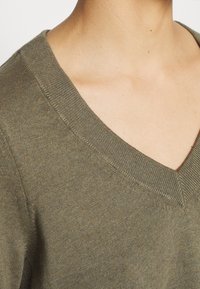 Gros plan d'une personne portant un pull en maille vert olive à col en V, montrant l'encolure et le haut de la poitrine.