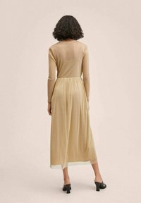 Robe longue, beige, avec des manches longues, un corsage ajusté et une jupe fluide. Fabriquée en matière transparente, elle présente une couture à la taille et un design à volants.