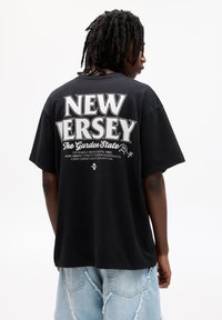 Sort bomulds t-shirt med "NEW JERSEY" i stort hvidt tekst på ryggen, sammen med beskrivende tekst i en mindre skrifttype.