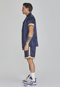 navy-blaues Kurzarmhemd und passende Shorts mit goldenen Akzenten; geometrisches Muster; kombiniert mit weißen Knöchelsocken und klobigen Turnschuhen.