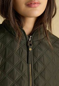 Olivový prešívaný kabát s vysokým golierom, predným zipsom so zlatou sponou a diamantovým vzorom na látke.
