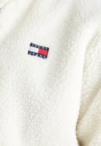 Krémová fleecová bunda s textúrovaným povrchom. Má obdĺžnikovú etiketu s nápisom "TOMMY JEANS" s červenými, bielymi a modrými akcentmi.