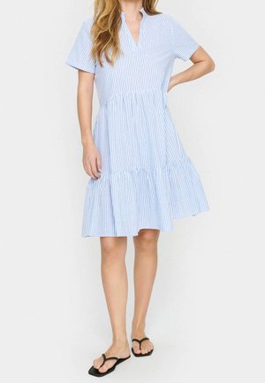 Mujer con vestido de manga corta a rayas verticales azules y blancas con bajo con volantes y sandalias negras tipo tanga, de pie con una mano en la cadera.