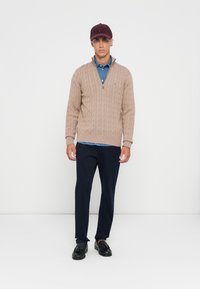 Jonge man in een beige kabelgebreide ziptrui over een blauwe shirt, marineblauwe broek, zwarte tassel loafers en een bordeauxrode pet op een witte achtergrond.