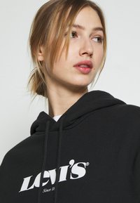 Felpa nera con cappuccio e logo bianco di Levi's, con cordini regolabili e texture morbida. Il design include una tasca a marsupio e polsini a costine.