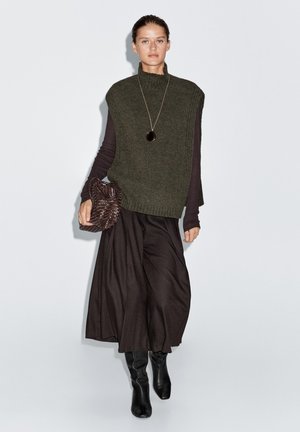 Gilet a maglia verde oliva con colletto alto sopra una maglietta scura a maniche lunghe, abbinato a una gonna nera fluida e stivali neri al ginocchio. Borsa marrone con texture.