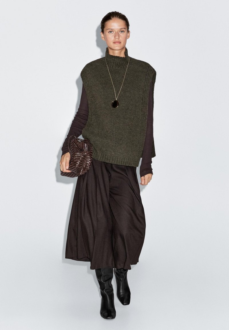 Olive green knit vest with a high collar over a dark long-sleeve top, paired with a flowy black skirt and knee-high black boots. Brown textured bag.