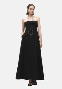 Svart, off-the-shoulder maxiklänning med figurnära överdel och utsvängd kjol, med sidofickor och ett stort cirkulärt hänge som halsband.