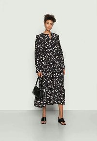 Vestido maxi preto com padrões brancos, mangas longas e gola com volantes. Combinado com uma bolsa preta e sandálias pretas de dedo.