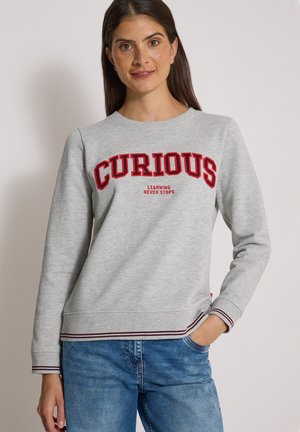 Femme portant un sweat-shirt gris clair avec un texte rouge "CURIOUS" et un texte plus petit "LEARNING NEVER STOPS,", associé à un jean bleu, main dans la poche.
