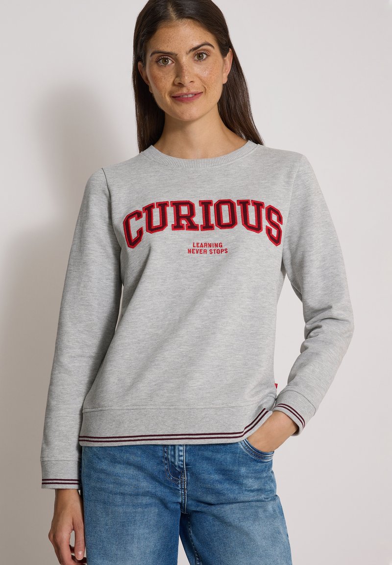 Femme portant un sweat-shirt gris clair avec un texte rouge "CURIOUS" et un texte plus petit "LEARNING NEVER STOPS,", associé à un jean bleu, main dans la poche.