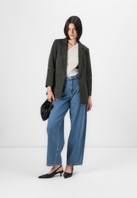 Una persona está de pie en un espacio blanco, vistiendo un blazer verde oliva, una blusa beige, jeans de pierna ancha y tacones negros con tiras, sosteniendo un bolso negro.