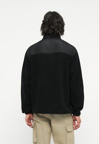 Schwarze Fleecejacke mit strukturiertem Oberteil und glattem Stoffeinsatz. Sie verfügt über einen hohen Kragen und eine lockere Passform, kombiniert mit hellbeigen Cargo-Hosen.