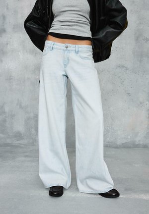 Persoană purtând blugi largi de culoare deschisă, pantofi negri, top gri scurt și jachetă de piele neagră, cu mâinile în buzunare.