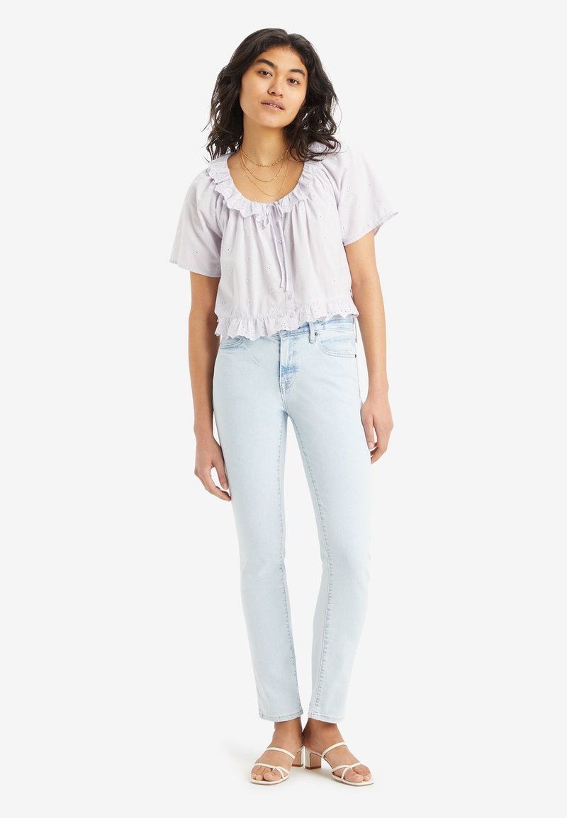 Hellblaues, kurzärmliges Blusenshirt mit rüschiertem Saum und Bindeband; kombiniert mit hellblauen, schmal geschnittenen Jeans und weißen, sandalenähnlichen Schuhen.
